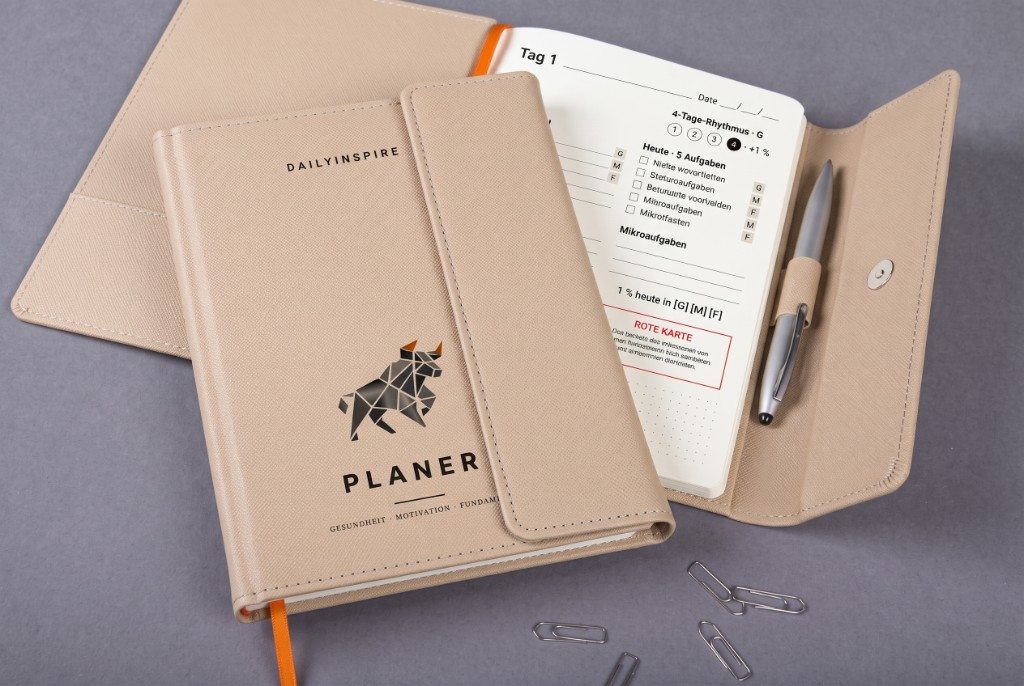 DailyInspire Planner, sand-beige A5 with flap, open on a day page, silver pen in the loop.
